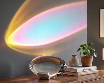 May include: A clear, oval-shaped glass lamp with a silver base labeled Prism Aura, projecting a vibrant, curved rainbow light effect onto a dark wall. The lamp sits on a wooden surface next to a small potted plant and two stacked books.