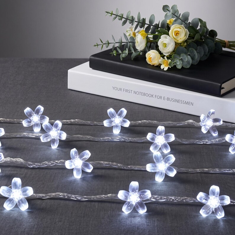 May include: A string of LED fairy lights featuring translucent, five-petaled flower-shaped covers that emit a bright white glow. The lights are connected by a thin, flexible clear wire, arranged in multiple rows across a dark surface.