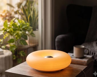 May include: A circular, donut-shaped table lamp with a smooth, matte, light-orange translucent shade that emits a warm glow. The center of the lamp features a small, metallic silver circular accent. A thin white power cord extends from the base.
