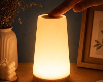 May include: A conical table lamp with a smooth, frosted white shade that emits a warm, soft glow. The lamp features a light-toned wooden base and a touch-sensitive top surface, providing a clean, functional design for ambient lighting.
