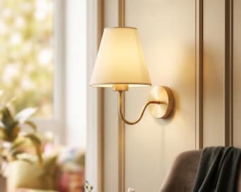 May include: A wall-mounted lamp featuring a brushed brass finish, a curved arm, and a tapered off-white fabric lampshade. The fixture is attached to a circular brass wall plate, emitting a warm, soft glow from the illuminated shade.