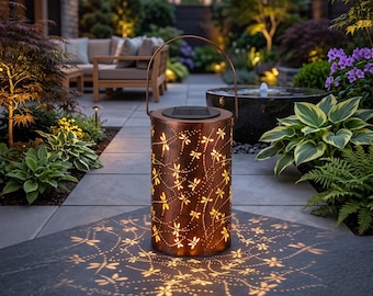 May include: A cylindrical copper-colored solar lantern with a metal handle and intricate dragonfly cutouts. The lantern emits a warm light, casting detailed dragonfly-shaped shadows onto the stone surface below, creating a decorative garden effect.