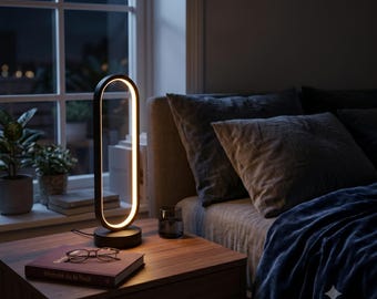 May include: A black oval-shaped LED table lamp with a continuous glowing light strip along the inner edge, mounted on a circular base. The lamp emits a warm light, illuminating the surrounding wooden surface and a book titled Serenite de la Nuit.