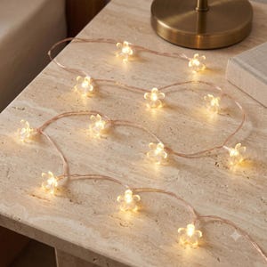 Puede incluir: Una cadena de luces LED con cubiertas translúcidas en forma de flor de cinco pétalos. Las luces emiten un brillo cálido y suave, conectadas por un cable fino y flexible de color cobre, creando una guirnalda decorativa delicada e iluminada.
