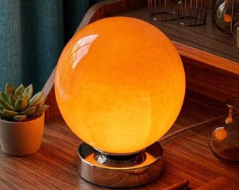 May include: A spherical orange glass table lamp with a smooth, glowing surface. The lamp is mounted on a circular, reflective silver-toned metal base. The light emits a warm, vibrant orange hue, illuminating the surrounding area with a soft glow.