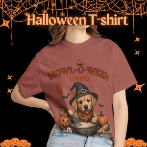 Può includere: Una t-shirt rosa polvere con la grafica di un cucciolo di golden retriever con un cappello da strega, che tiene una frusta sopra una ciotola. Il testo recita "HOWL-O-WEEN treats" con una zucca sopra, pipistrelli e un mattarello.