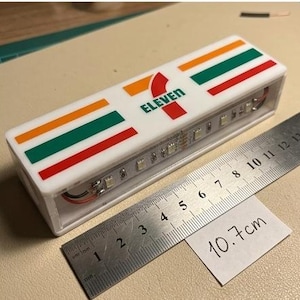 May include: A rectangular white plastic sign featuring the 7-Eleven logo with orange, green, and red stripes. The sign has an open side revealing an internal LED light strip. A metal ruler is placed alongside, showing a length of 10.7 cm.