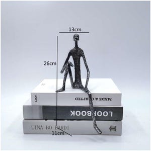 Pode incluir: Uma escultura de metal cinza-escuro texturizado de uma figura sentada com membros alongados. A figura está posicionada sobre uma pilha de três livros. As dimensões marcadas na imagem são 13 cm de largura, 26 cm de altura e 11 cm de profundidade.
