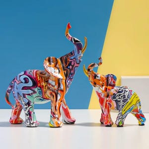 Peut inclure: Deux figurines d'éléphants côte à côte, arborant un motif graffiti vibrant avec des éclaboussures d'orange, de bleu, de jaune et de noir. Les deux sculptures représentent des éléphants avec la trompe levée, affichant une finition brillante.