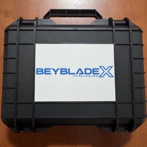 Può includere: Una valigetta rigida rettangolare nera con una maniglia superiore e un'etichetta rettangolare bianca sulla parte anteriore. L'etichetta presenta il testo blu BEYBLADE X con caratteri giapponesi sotto la parola BEYBLADE.