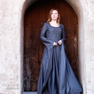 May include: A woman in a long, dark gray dress stands in a doorway. The dress has a round neckline and long sleeves. The woman is looking up and has long, red hair.