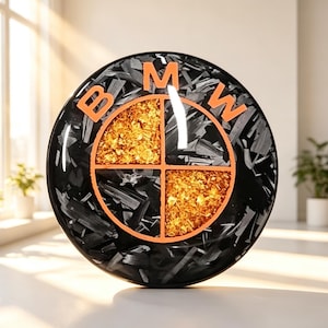 May include: A circular BMW emblem featuring a glossy finish with a black and gray carbon fiber texture. The logo is divided into four quadrants, with two sections filled with gold-colored flakes and two with dark carbon fiber. The letters BMW are orange.