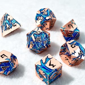 Könnte beinhalten: Ein Set aus sieben polyedrischen Würfeln mit einem metallischen Roségold-Finish und vertieften Bereichen, die mit leuchtend blauem und türkisfarbenem Emaille gefüllt sind. Jede Seite zeigt schwarz gravierte Zahlen in einer kantigen Runenschrift.