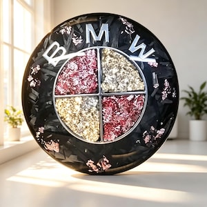 May include: A circular BMW emblem featuring a black background with embedded dark carbon fiber-like textures and scattered rose gold foil flakes. The central logo is divided into four quadrants filled with alternating rose gold and gold metallic flakes.