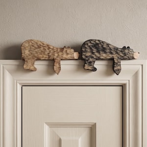 May include: Two carved wooden bear figures resting on top of a door frame. One bear is light brown with a natural wood grain, and the other is dark brown with a charred finish. Both figures feature textured surfaces and simple, stylized shapes.