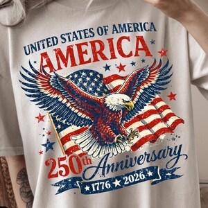 May include: A beige t-shirt featuring a graphic of a bald eagle with wings spread in front of an American flag. The text reads UNITED STATES OF AMERICA, AMERICA, 250th Anniversary, and 1776-2026. Red and blue stars are scattered across the design.