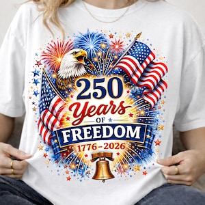 May include: A white t-shirt featuring a graphic of a bald eagle, two American flags, and fireworks. The text reads 250 Years OF FREEDOM 1776-2026, accompanied by an illustration of the Liberty Bell centered at the bottom of the patriotic design.