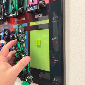 Puede incluir: Un marco que muestra coches de carreras de Fórmula 1 en miniatura dispuestos en filas con nombres de equipos como Williams y Kick Sauber. Una sección rectangular de color verde brillante incluye un pequeño bloque de construcción verde lima.
