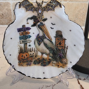 May include: A white scallop shell dish featuring a glittery pelican wearing a witch hat and flip-flops. The design includes pumpkins, a signpost reading "To the Beach Boo!", and a "Happy Halloween" banner. A jeweled bat ornament sits at the top.