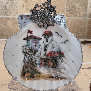 May include: A decorative seashell featuring a glittery ghost dressed as a lifeguard, wearing a red cap and holding a red rescue buoy. The scene includes a beach patrol tower with the text 'BEACH PATROL' and a sign reading 'SWIM AT YOUR OWN RISK'.