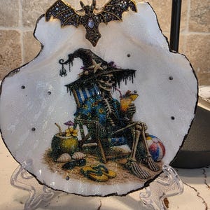 May include: A decorative seashell dish featuring a glittery illustration of a skeleton wearing a witch hat and floral shirt, sitting in a beach chair with a cocktail. A gold-toned bat ornament is attached to the top edge of the white shell.