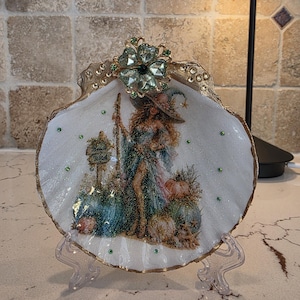 May include: A decorative scallop shell featuring a glittery illustration of a witch standing near pumpkins and a sign that reads Beach Witch. The shell is accented with gold paint on the edges, small green rhinestones, and a large floral crystal topper.