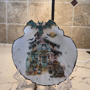 May include: A decorative scallop shell featuring a glittery white finish and a central illustration of a haunted house with ghosts, pumpkins, and a skeleton. A teal jeweled bat is attached to the top edge, and the house displays the text BEWARE.