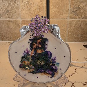 May include: A decorative seashell dish featuring a glittery mermaid illustration with purple and green accents, adorned with a large purple rhinestone flower brooch at the top. The shell has a silver-painted edge and small purple rhinestones.
