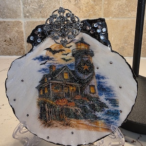 May include: A decorative oyster shell featuring a decoupage scene of a lighthouse and house at night with pumpkins, bats, and a full moon. The top edge is painted black and adorned with a silver-toned filigree brooch and clear rhinestones.