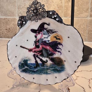 May include: A decorative seashell featuring a painted illustration of a pink flamingo dressed as a witch, flying on a broomstick over ocean waves with a full moon and bats in the background. The shell is adorned with clear rhinestones and a metal filigree.