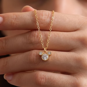 Peut inclure: Un collier doré avec un pendentif en forme de silhouette de tête de souris. Le pendentif est composé de trois pierres rondes transparentes serties dans du métal doré, avec une pierre centrale plus grande et deux plus petites au-dessus.