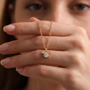 Peut inclure: Un collier doré avec un pendentif en forme de silhouette de tête de souris. Le pendentif est composé de trois pierres rondes transparentes serties dans du métal doré, avec une pierre centrale plus grande et deux plus petites au-dessus.