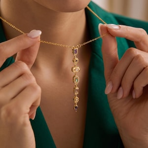 May include: A gold-tone chain necklace featuring a vertical drop pendant composed of seven distinct geometric links. Each link is set with a different colored gemstone, including purple, black, clear, and teal stones, arranged in a linear sequence.