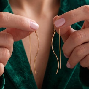 May include: A pair of gold-toned threader earrings featuring a long, slender straight post connected to a delicate, gently curved wave-shaped wire. The earrings are crafted from a thin, polished metal, creating a minimalist and elongated silhouette.