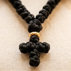 Pode incluir: Um colar de cordão preto com nós, apresentando um pingente de cruz feito do mesmo cordão preto. Um anel de metal dourado liso e polido separa a cruz da corrente principal do colar, que consiste em uma série de nós redondos repetidos.