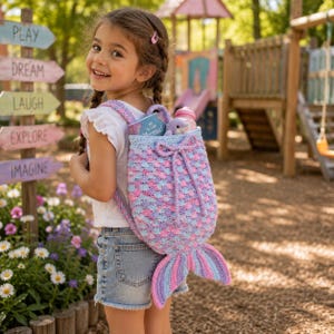 May include: A crochet backpack shaped like a mermaid tail in shades of pink, purple, and blue. The bag features a drawstring closure with a bow, a textured scale pattern, and a prominent tail fin at the bottom. A small book and bottle are inside.