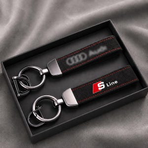 Puede incluir: Dos llaveros de ante negro con herrajes metálicos y anillas. Un llavero presenta el logotipo de Audi, mientras que el otro muestra el emblema S Line en rojo, blanco y negro. Ambas correas tienen costuras rojas a lo largo de los bordes.