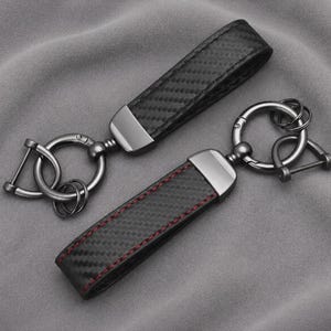 May include: Two black keychains featuring a carbon fibre pattern and dark metallic hardware. Each keychain includes a rectangular strap attached to a circular ring and a D-shaped shackle. One strap displays red stitching along the edges.