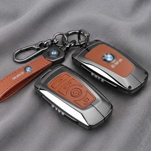 Peut inclure: Deux clés de voiture BMW logées dans des coques en métal gris foncé brillant avec des inserts en cuir beige, présentant des icônes de boutons en relief. Une lanière de porte-clés en cuir beige assortie avec un clip en métal gris foncé est incluse.
