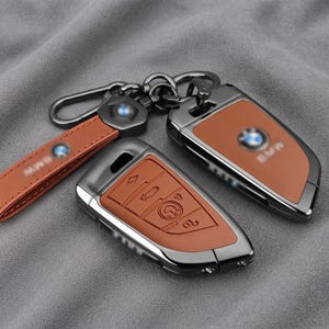 Peut inclure: Un ensemble de coques pour clé de voiture BMW composé d'un cadre gris métallique foncé avec des inserts en cuir marron clair. L'ensemble comprend un étui pour clé avec des icônes de boutons en relief, une coque de protection et un porte-clés.