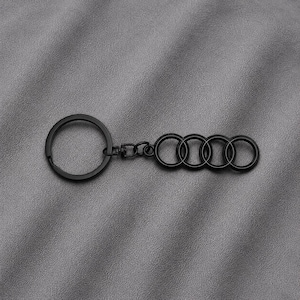 May include: A matte black metal keyring featuring a circular split ring connected by a small swivel link to a horizontal emblem consisting of four interlocking rings. The design is uniform in colour and finish, presenting a sleek, solid construction.
