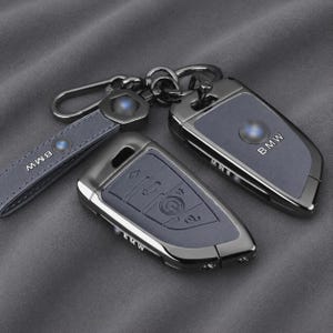 Peut inclure: Un ensemble de coques de clé de voiture BMW en métal gris foncé, accompagné d'une lanière en cuir assortie avec un clip en métal. La coque présente une finition lisse avec le logo BMW gravé et inclut une protection pour les boutons.