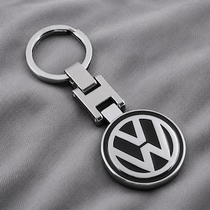 Könnte beinhalten: Ein silberfarbener Metallschlüsselanhänger mit dem kreisförmigen Volkswagen-Logo auf schwarzem Hintergrund. Das Design umfasst ein bewegliches Verbindungsstück, das an einem runden Standard-Schlüsselring befestigt ist, alles in polierter Optik.