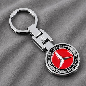 Könnte beinhalten: Ein silberfarbener Metallschlüsselanhänger mit einem kreisförmigen Emblem, das ein rotes Zentrum und das Mercedes-Benz-Logo aufweist. Das Design umfasst einen silbernen dreizackigen Stern, schwarze Schrift und einen Lorbeerkranz.