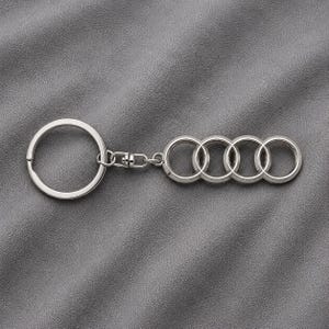 Peut inclure: Un porte-clés en métal argenté composé d'un anneau brisé circulaire relié par un petit maillon pivotant à une breloque formée de quatre anneaux entrelacés disposés sur une ligne horizontale, rappelant un logo automobile classique.