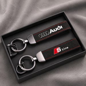 Può includere: Due portachiavi in camoscio nero con cuciture rosse e componenti in metallo argentato. Un portachiavi presenta il logo e la scritta Audi in bianco, l'altro l'emblema S Line in rosso e bianco. Entrambi includono anelli metallici circolari.
