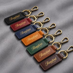 May include: A collection of seven rectangular keychains in black, red, blue, orange, green, light brown, and dark brown. Each keychain features a gold-toned metal lobster clasp and ring, with the word Rangland embossed in gold script lettering.