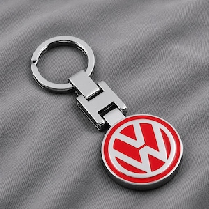Könnte beinhalten: Ein silberfarbener Metallschlüsselanhänger mit einem runden Anhänger, der einen roten Hintergrund und das weiße Volkswagen-Logo aufweist. Der Schlüsselanhänger besitzt ein rechteckiges Gelenkstück an einem Standard-Schlüsselring.