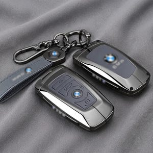 Peut inclure: Deux télécommandes de voiture gris métallisé foncé avec une finition brillante et une lanière de porte-clés en cuir assortie. Les boîtiers présentent un logo BMW circulaire central et des boutons intégrés. Le porte-clés inclut un clip en métal.