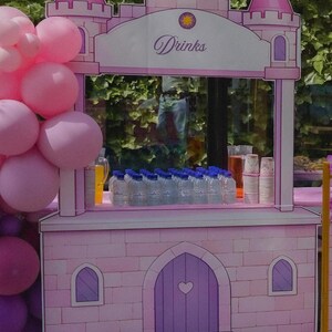 Op de afbeelding: Een roze drankstand in kasteelstijl met een centraal bord met het woord Drinks, een paarse boogdeur met een hartvormige uitsparing en twee zijtorens. De stand is versierd met roze ballonnen en bevat meerdere doorzichtige plastic waterflesjes.